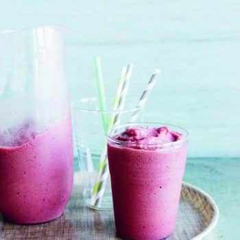 purple smoothie in a glass next to a pitcher with a blue counter and backsplash