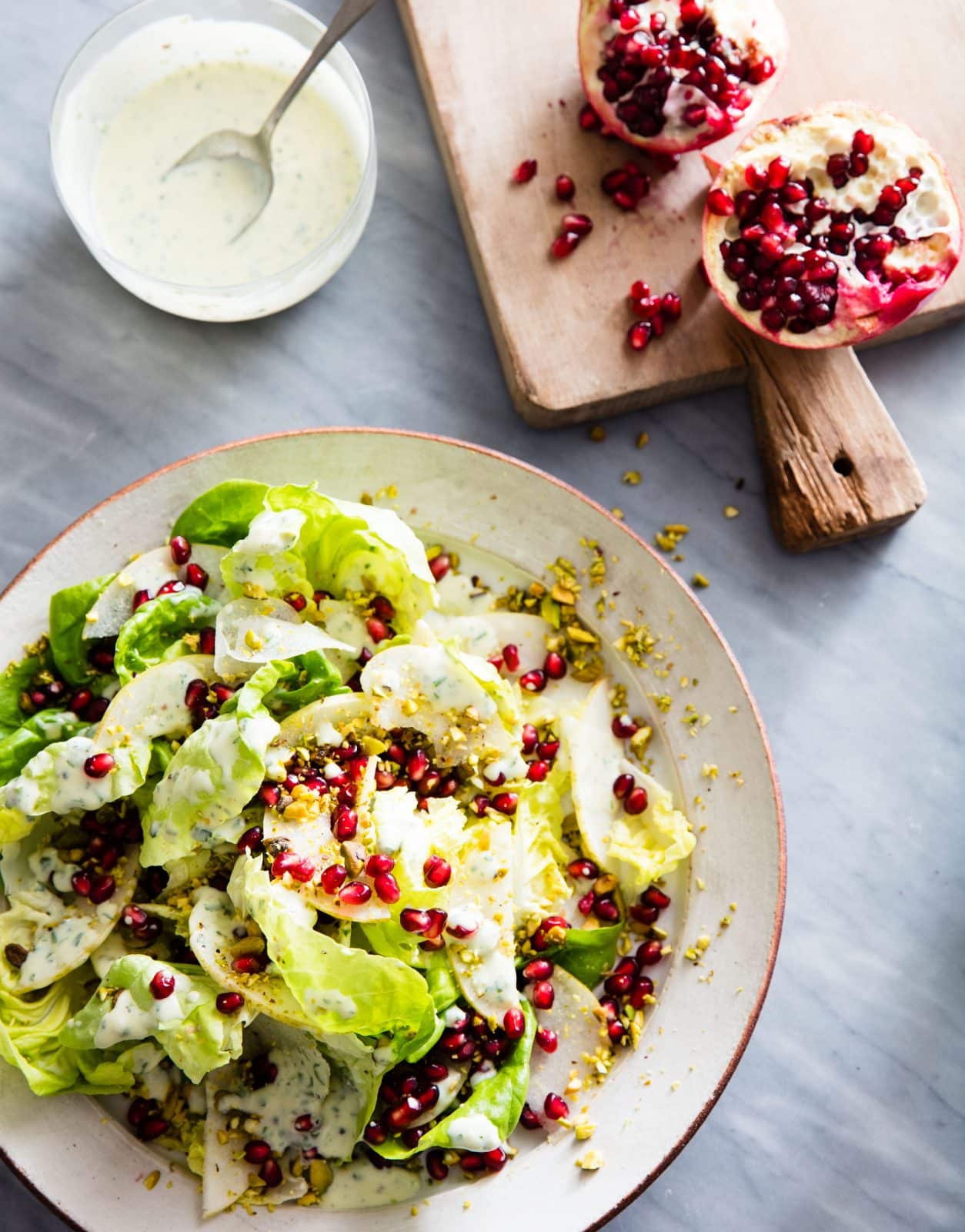butter lettuce salad next to pomegranates on a wood board and a white sauce with a spoon