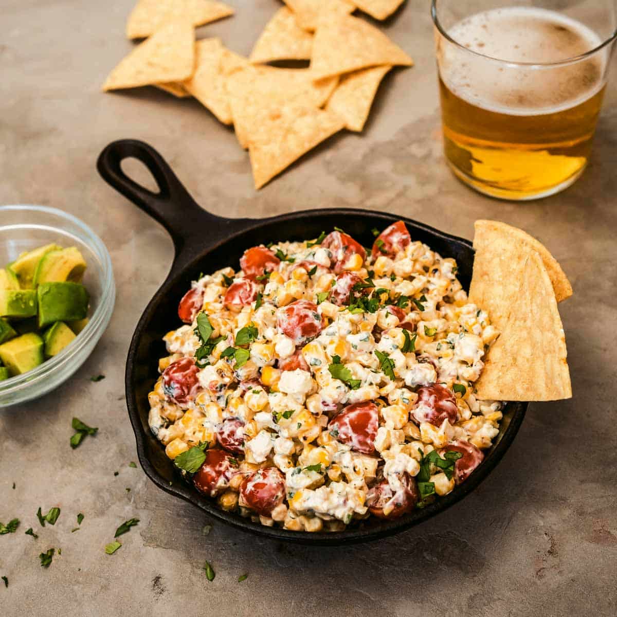 Corn dip in a cast iron skillet next to a glass of beer, chips, and avocado.