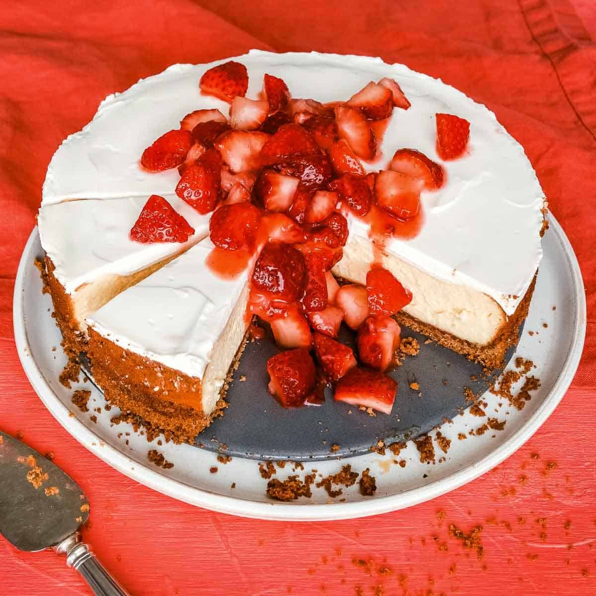 Vanilla cheesecake topped with chopped strawberries on a white platter on an orange table next to a spatula.