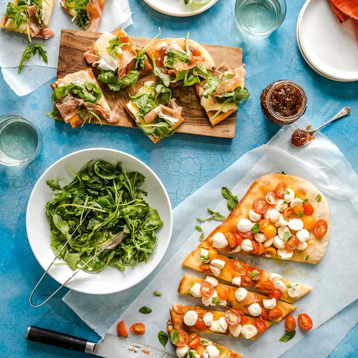 Proscuitto arugula flatbread and caprese flatbread sliced on a blue counter next to extra fig jam and arugula.