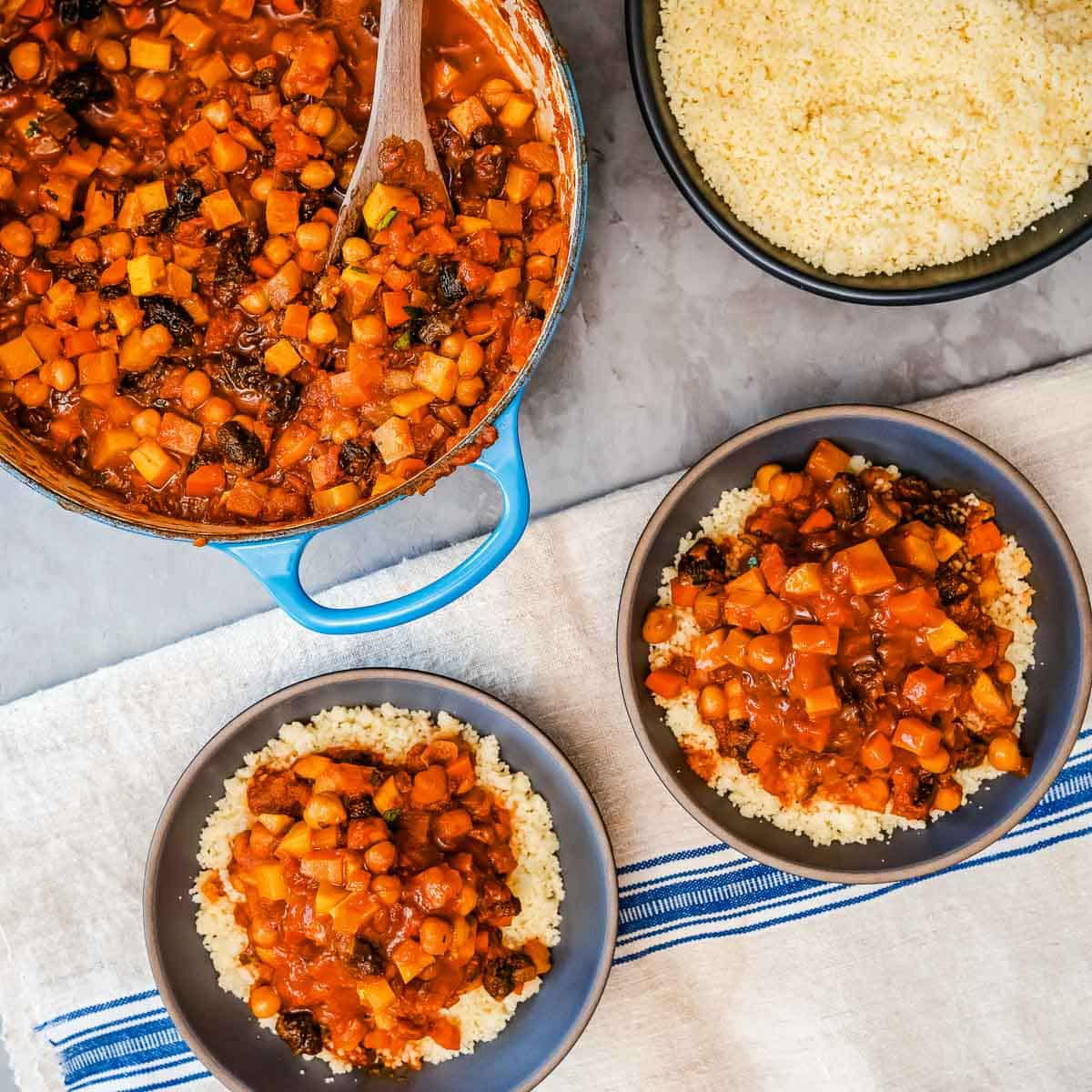 Two bowls of couscous topped with moroccan spiced root vegetables and beans next to an extra bowl of couscous and a Dutch oven of vegetables.